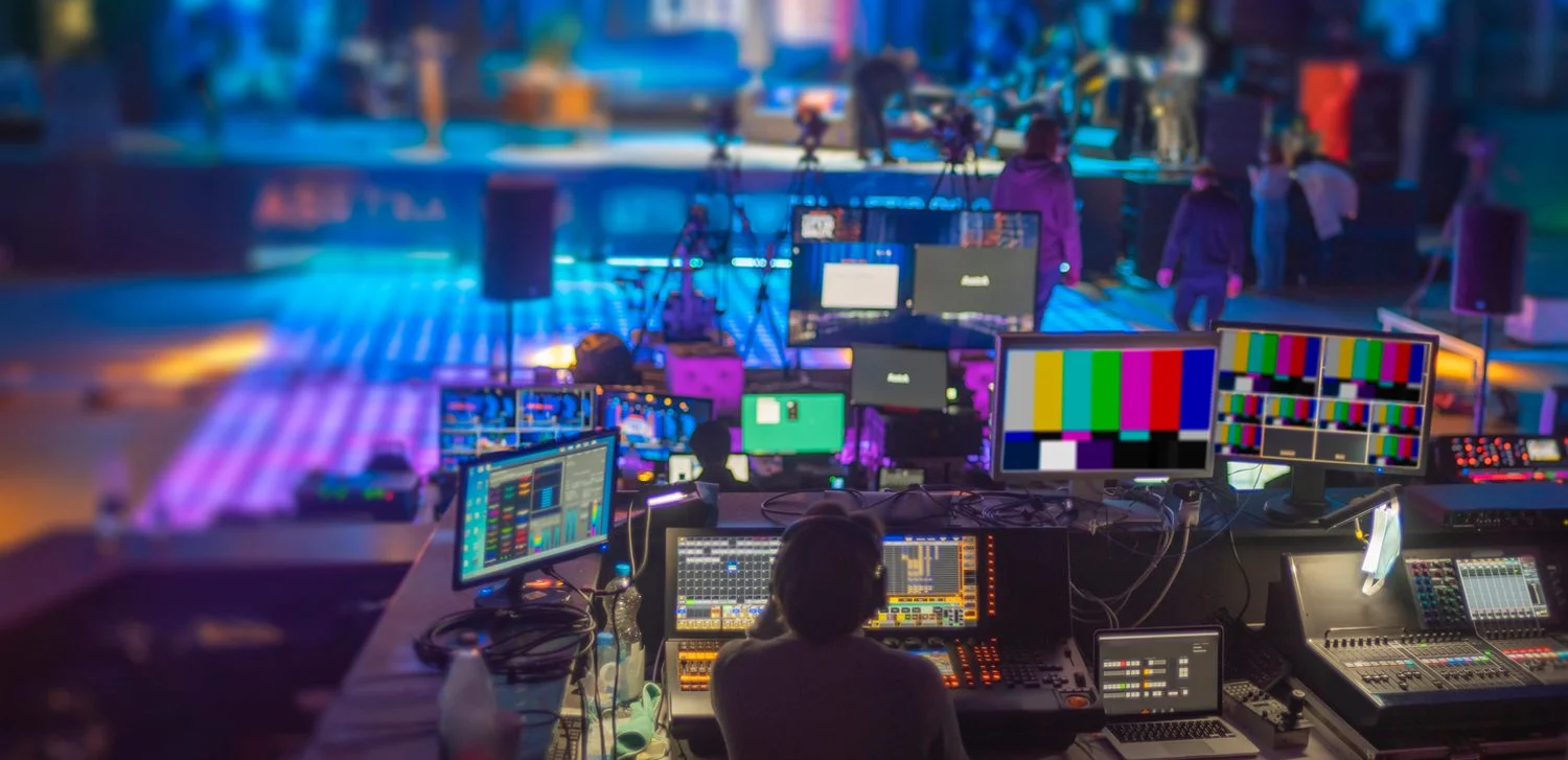 A live Remote production setup with multiple monitors, a mixing console, and cameras on tripods covering an event with proper lighting and crew members working.