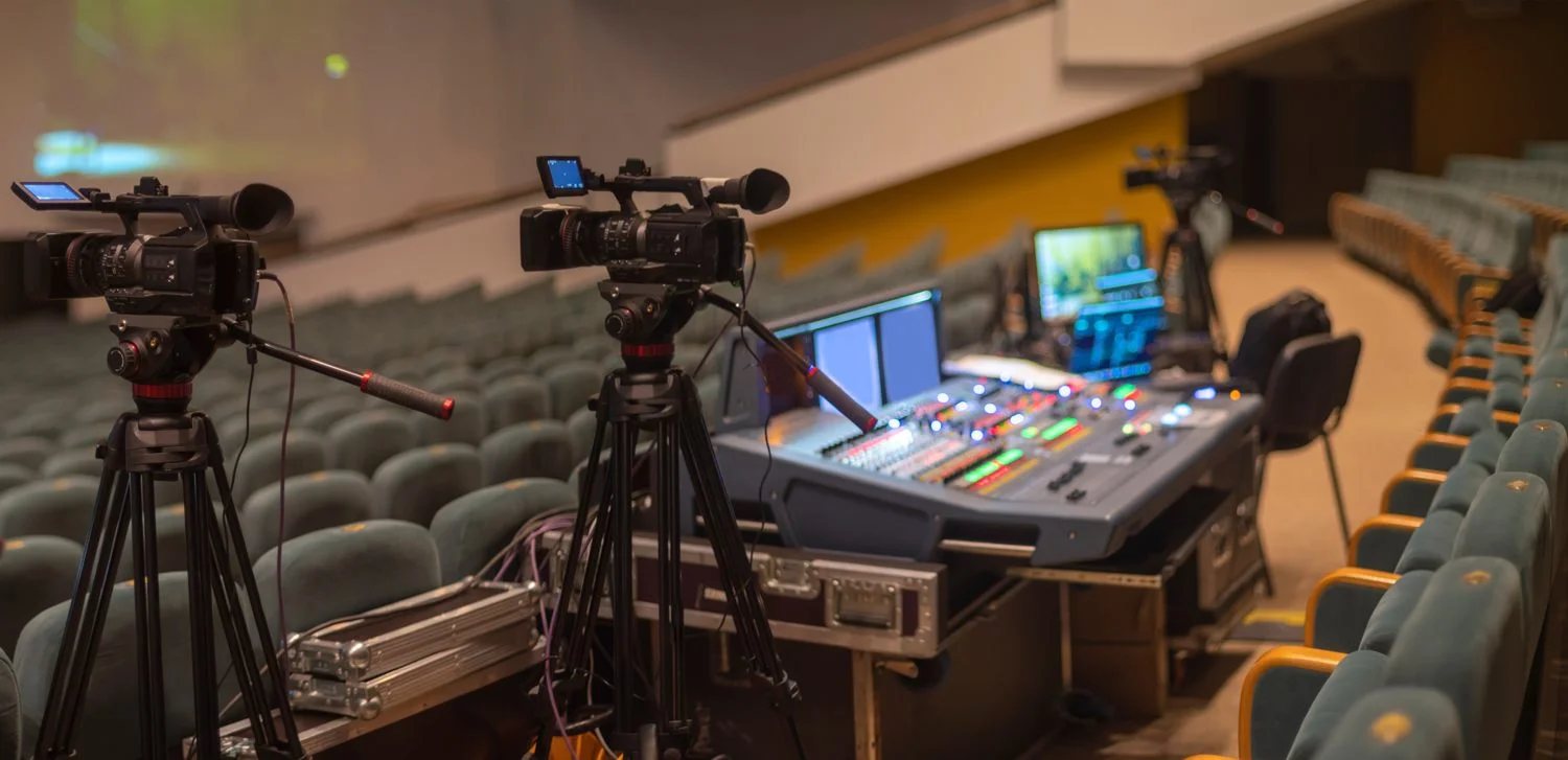 A live broadcast production setup with two cameras on a tripod, video switcher, preview and program monitor.